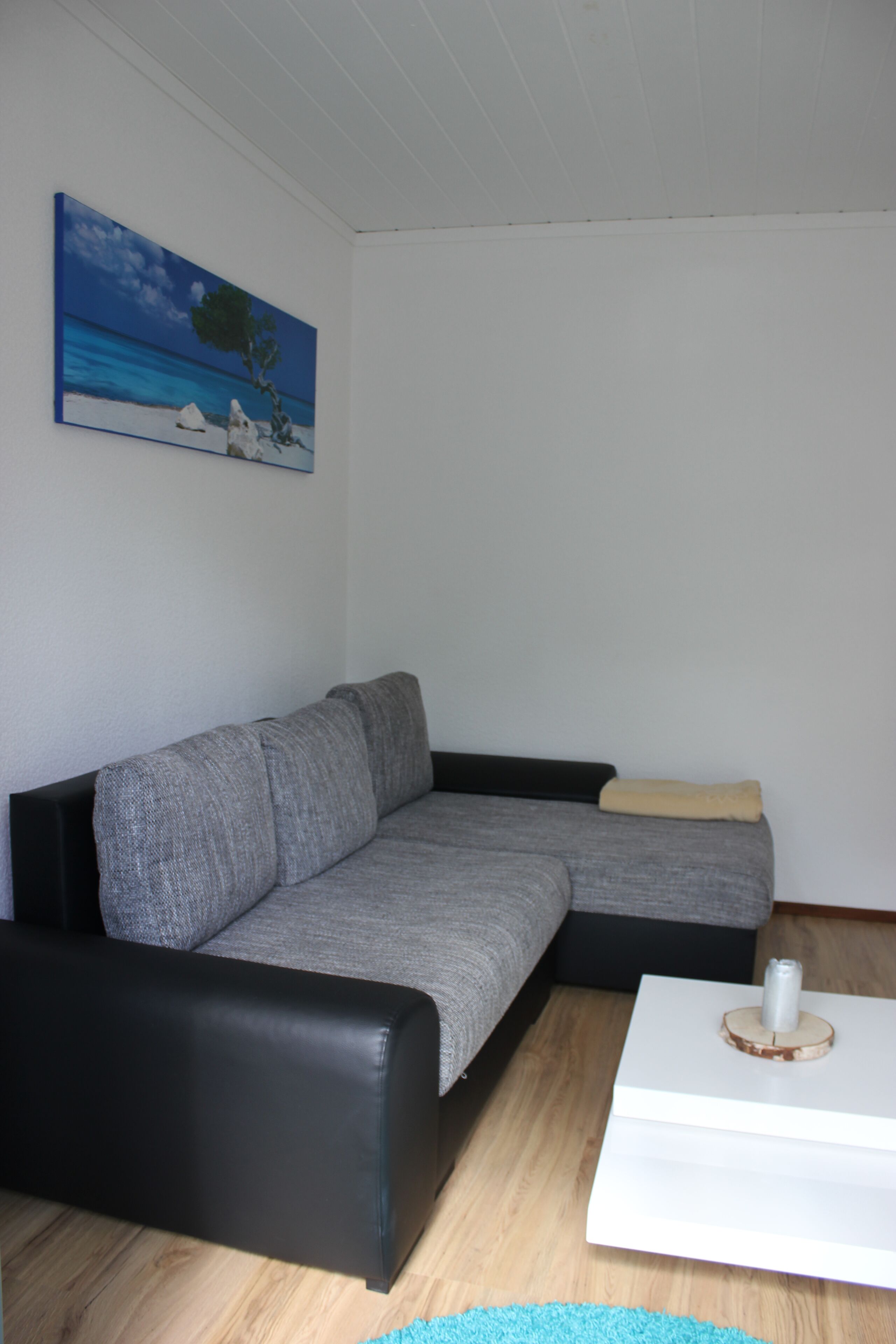 Un salón con un sofá seccional gris, una mesa de centro blanca y un cuadro con una playa en la pared.