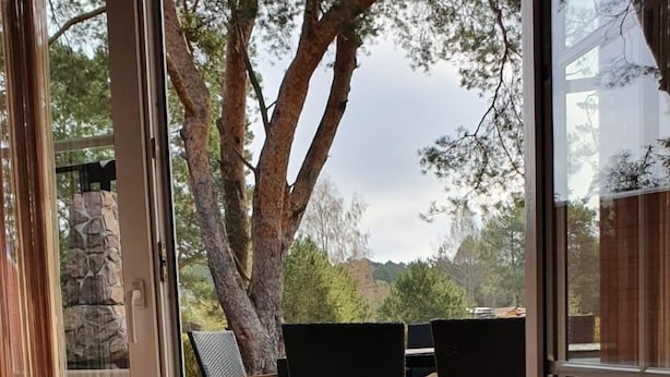 Eine offene Schiebetür führt zu einer Terrasse im Freien mit einer Steinmauer, einem großen Baum und einem Tisch mit Stühlen.