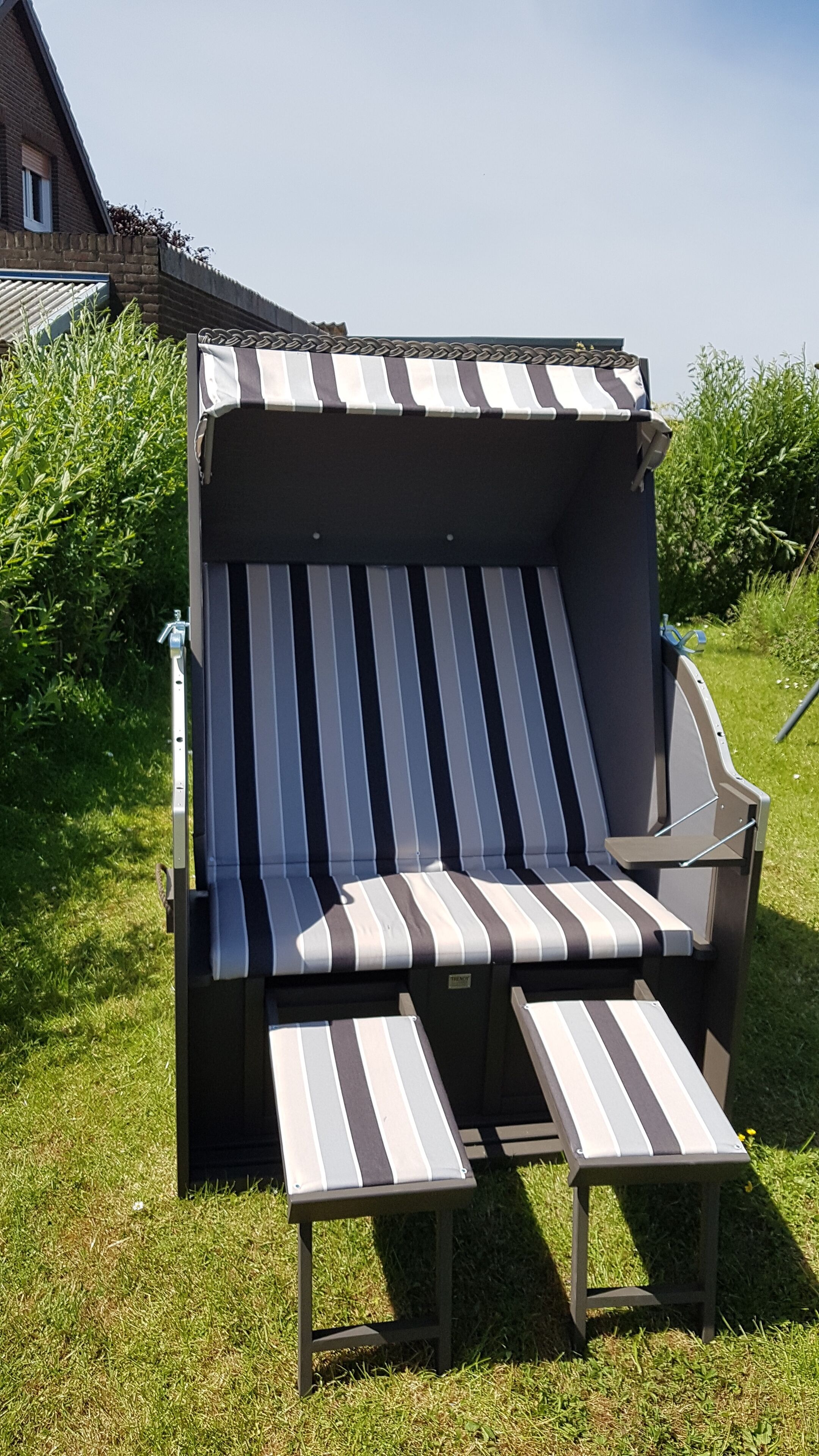 En strandstol med en stribet pude, placeret på græs med et hus og grøn vegetation i baggrunden.