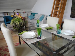 A dining area with a glass table set for four, surrounded by white chairs. A sofa with blue cushions is in the background, and a ladder leans against the wall. - Studio apartment in a dreamy and quiet garden in the immediate vicinity of the lake (Schondorf am Ammersee)