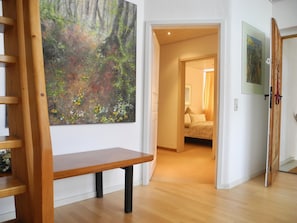 A room with a wooden bench, a painting of a forest, a bed with a white headboard, and a door leading to another room. - Studio apartment in a dreamy and quiet garden in the immediate vicinity of the lake (Schondorf am Ammersee)