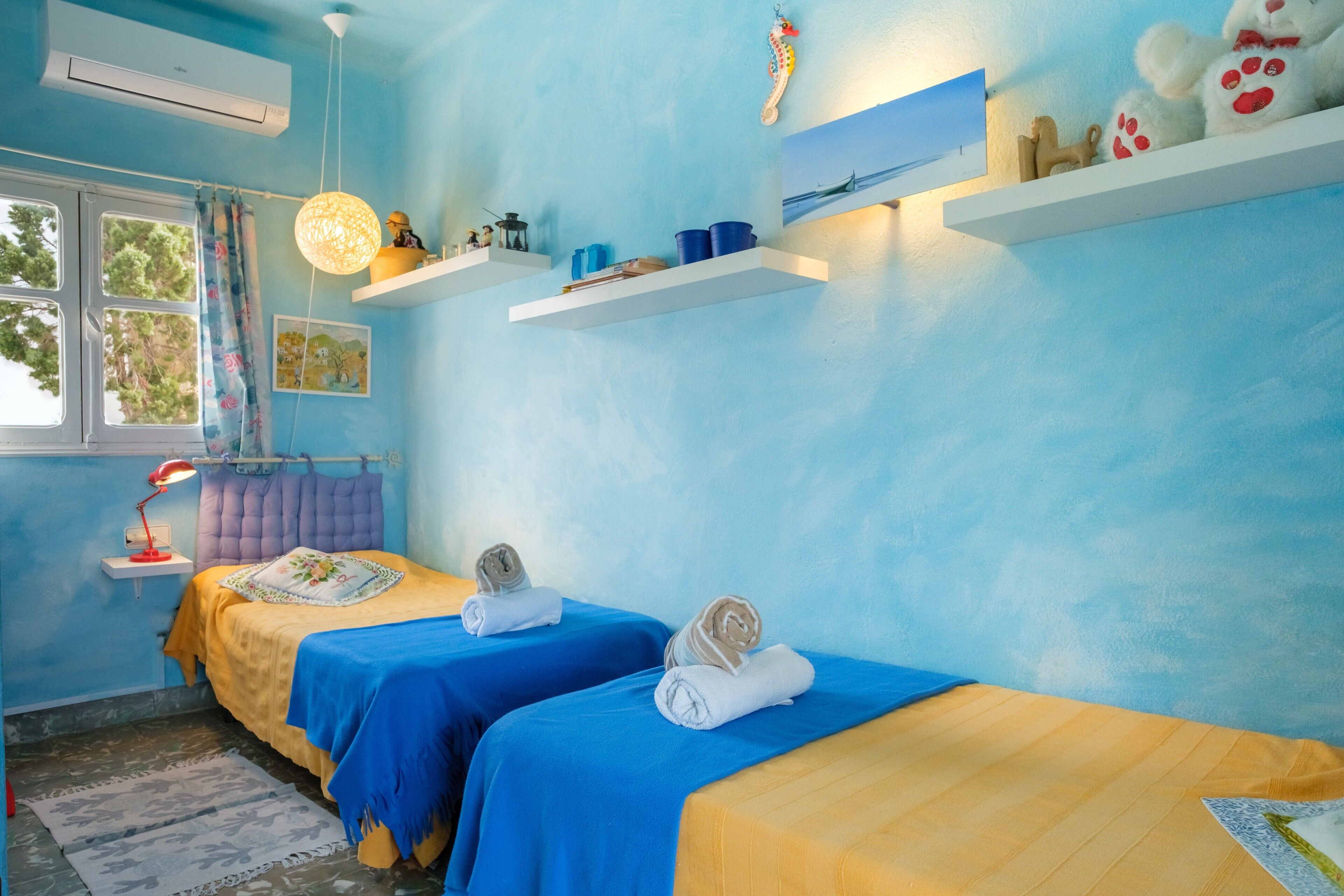 A room with two beds, a window, a hanging light fixture, and a shelf with various items.