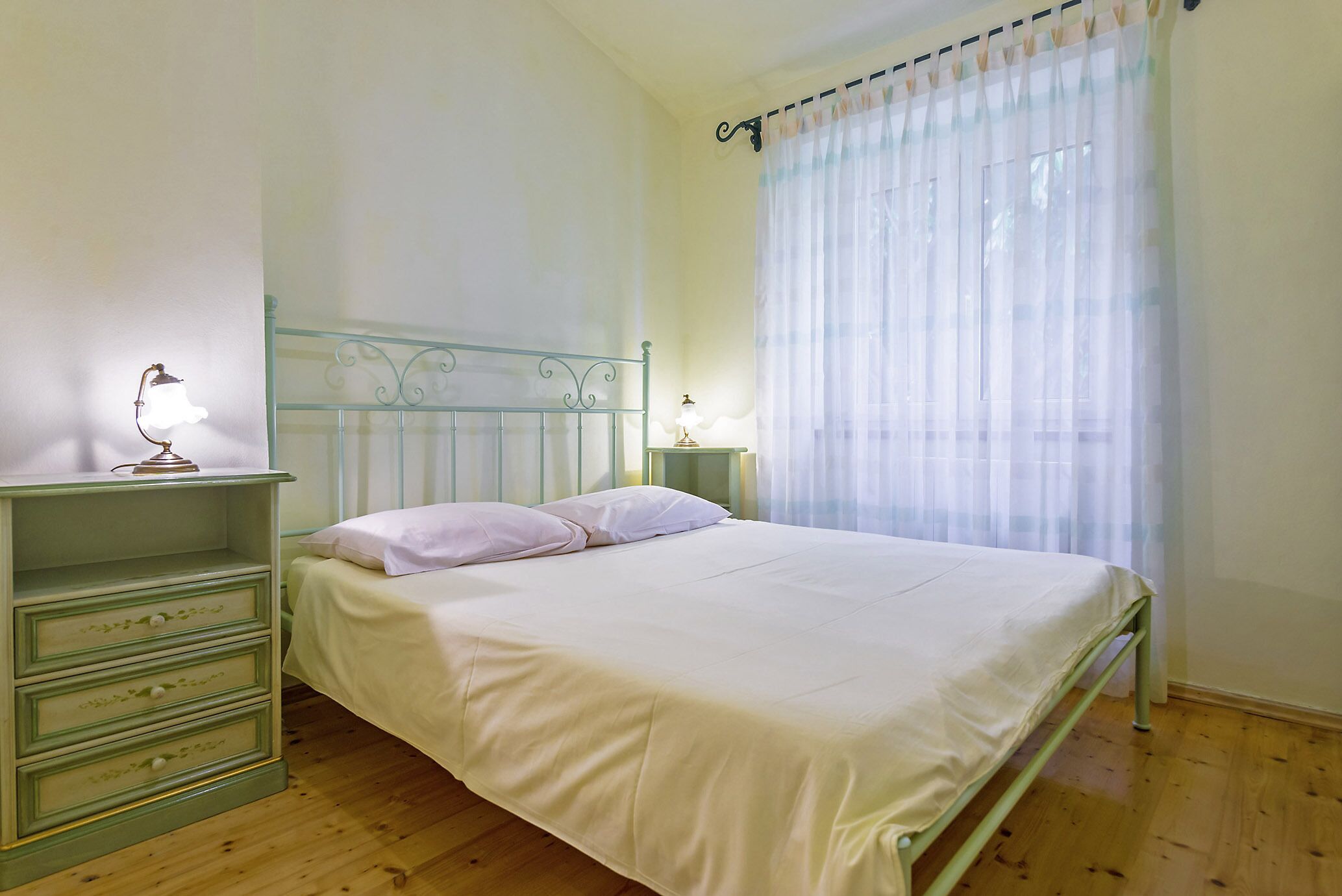 A bedroom with a single bed, a nightstand with a lamp, a dresser with drawers, and a window with sheer curtains.