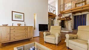 A spacious living room with a wooden floor, a glass coffee table, two beige armchairs, a wooden cabinet, a flat-screen TV, and a stone fireplace.