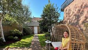 Un patio con una silla de mimbre, una pequeña mesa y un sendero de ladrillos que conduce a un edificio con techo de tejas.
