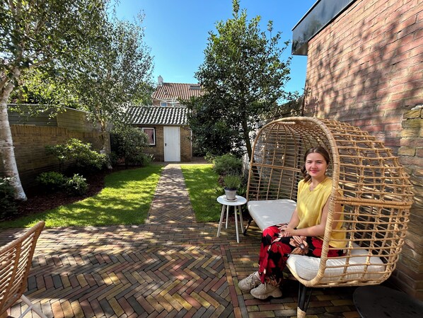 A patio area with a wicker chair, a small table, and a brick pathway leading to a building with a tiled roof.