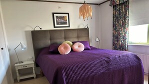 Un dormitorio con una cama grande, una mesita de noche, una lámpara, un cuadro en la pared y una ventana con cortinas.