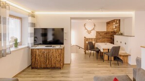 Ein modernes Wohnzimmer mit Couchtisch aus Holz, einem Flachbildfernseher, einem Essbereich mit Holztisch und Stühlen sowie einer großen Wanddekoration mit einem Hirschkopf.