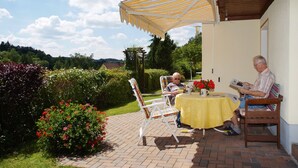Two people seated at a table under a striped canopy in an outdoor patio area with brick flooring and a garden with red flowers. - Apartment for 2-3 people on the southern slope with View and possibility of excursions (Plankenfels)