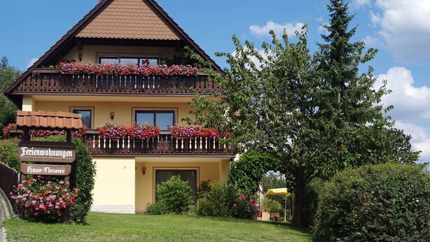 A two-story building with a balcony adorned with flowers, a sign indicating 'Ferienswohnungen Haus-Neuner', and a lush green garden. - Apartment for 2-3 people on the southern slope with View and possibility of excursions (Plankenfels)