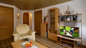 A cozy living room with a beige sofa, wooden entertainment unit with speakers, a television, and a table with fruit. - Apartment for 2-3 people on the southern slope with View and possibility of excursions (Plankenfels)