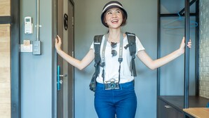 A person standing in a hallway with a door on the left, a coat rack on the right, and a small table with a chair. - Ray Hostel (Chiang Mai)
