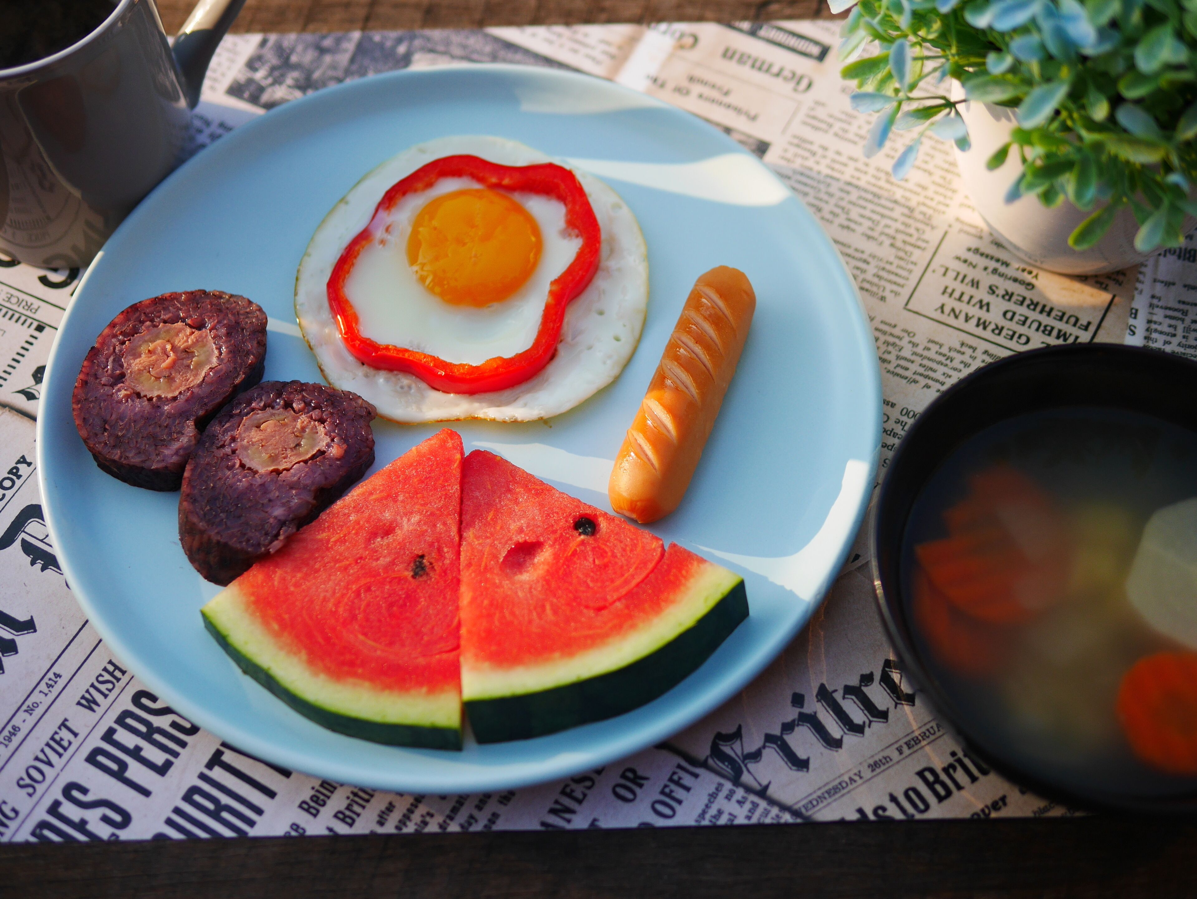 A plate with a sunny side up egg, sausage, and watermelon slices on a newspaper-covered table.