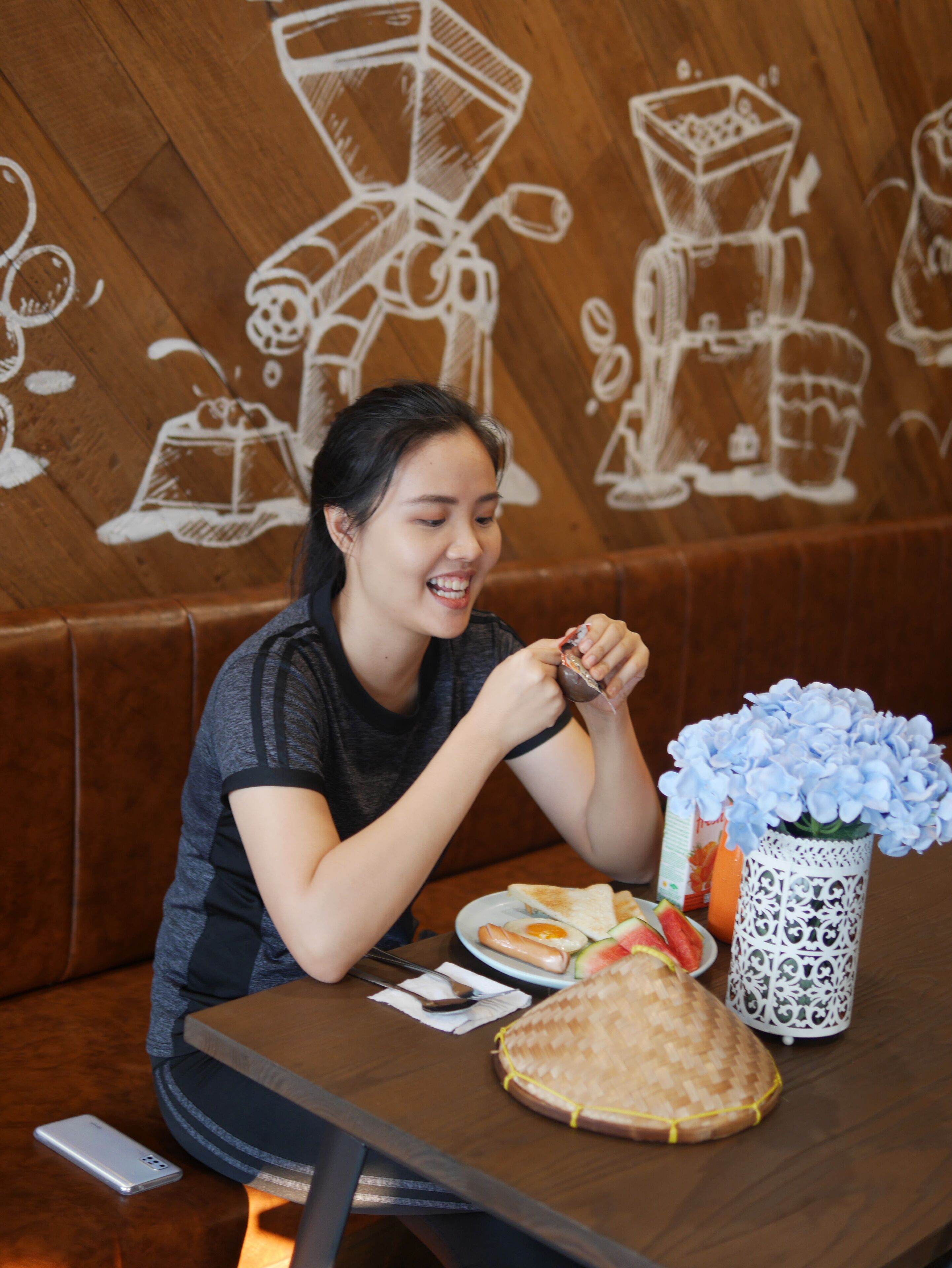 Una persona sentada a una mesa con comida, un teléfono inteligente y un jarrón decorativo con flores azules.