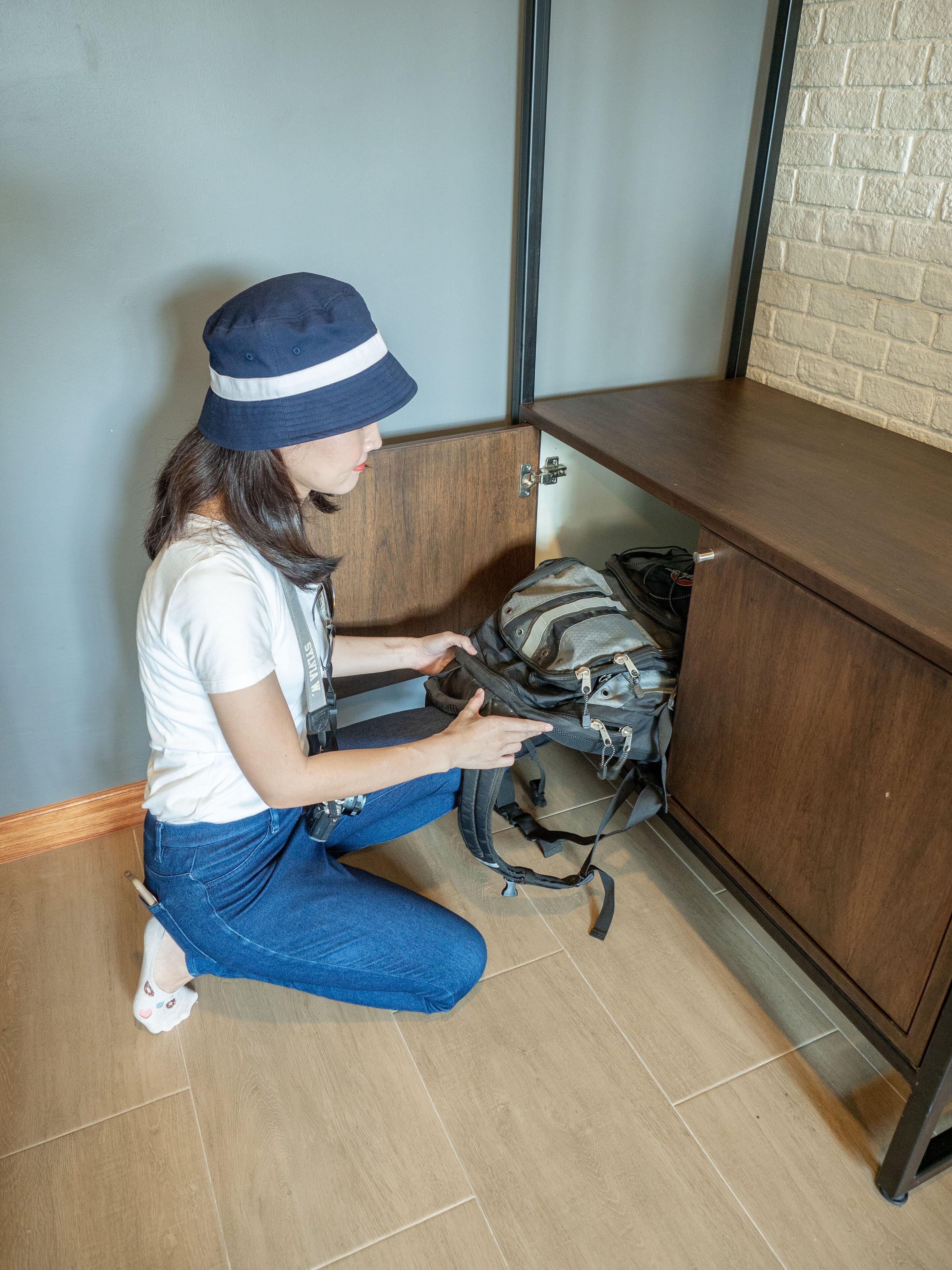 A person kneeling on a wooden floor, interacting with a backpack and a wooden cabinet.
