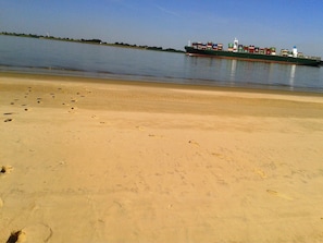 Ein sandiger Strand mit Fußspuren, die zum Wasser führen. Ein Frachtschiff ist in der Ferne sichtbar.
