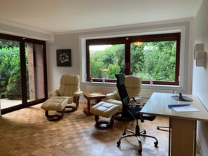 Ein geräumiges Zimmer mit großem Fenster und Blick auf Grünflächen, Holzboden und Schreibtisch mit Bürostühlen.