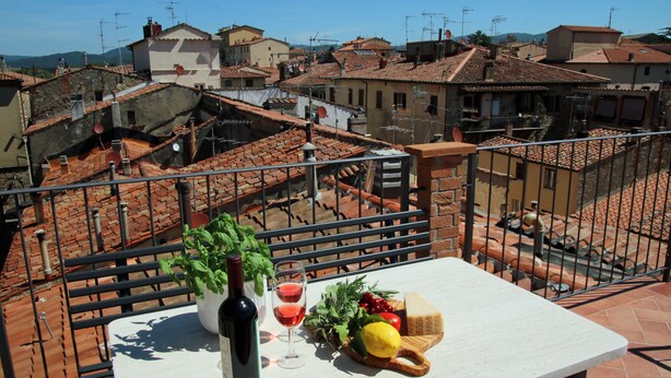 Un balcón con una mesa dispuesta para una comida, incluyendo una botella de vino, copas y verduras frescas.