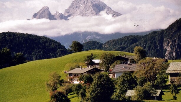 A rural house with a sloped roof, surrounded by lush green fields and trees, with a mountain range in the background.
