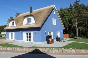 A blue house with a thatched roof, white shutters, and a small outdoor seating area.
