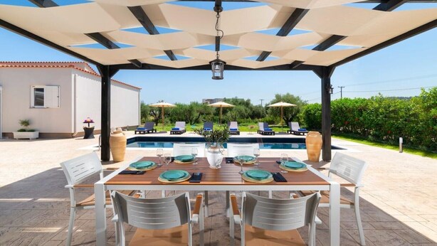 An outdoor dining area with a pool in the background, covered patio, and outdoor seating. - The idyllic oasis of well-being (Soroni)