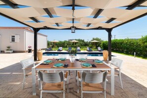 An outdoor dining area with a pool in the background, covered patio, and outdoor seating.