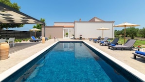 A rectangular swimming pool with lounge chairs and umbrellas on the deck, a covered seating area, and a building with a tiled roof in the background. - The idyllic oasis of well-being (Soroni)
