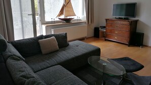 Een woonkamer met een bank, een glazen salontafel, een televisie, een dressoir en een model zeilboot bij het raam.
