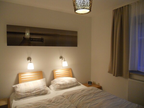 A hotel room with a bed, two bedside lamps, a nightstand, a wall-mounted artwork, and a window with curtains. - First floor apartment by the water with terrace, carport parking space and WLAN (Greifswald OT: Insel Riems)