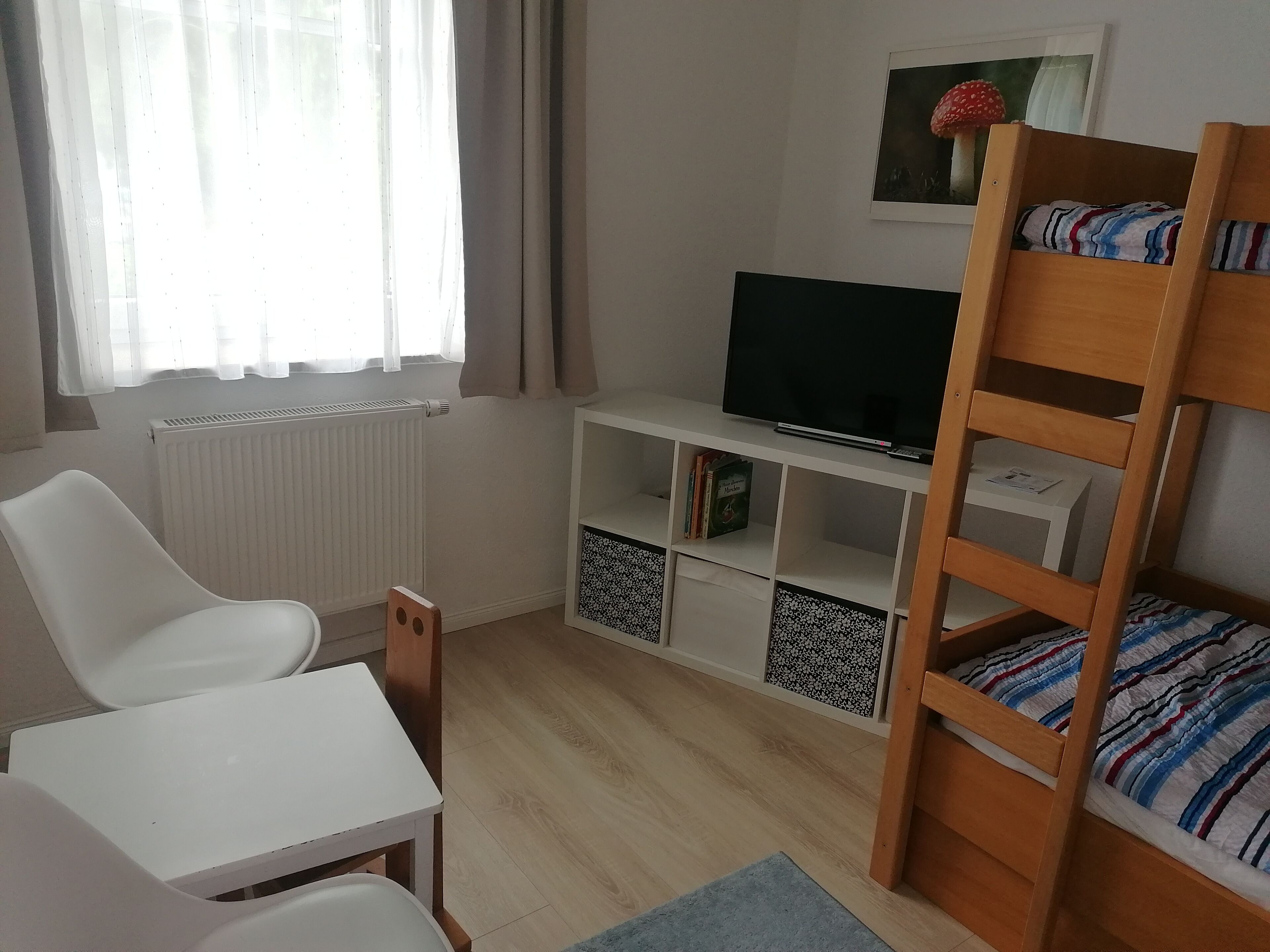 Une chambre avec un lit superposé, un bureau, une chaise, un téléviseur posé sur une étagère blanche et un radiateur.