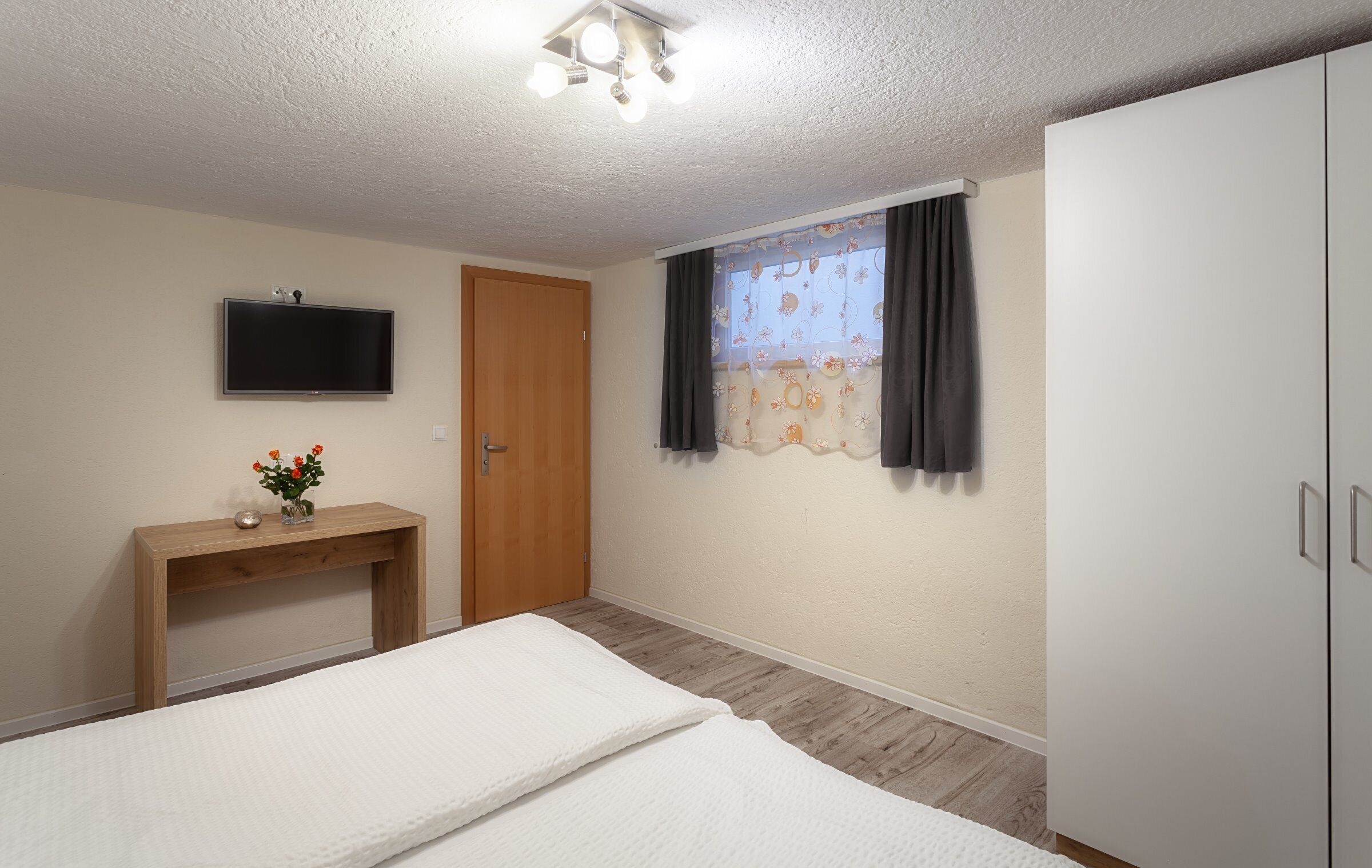 Een slaapkamer met een bed, een aan de muur bevestigde televisie, een houten nachtkastje met een vaas, een deur en een raam met gordijnen.