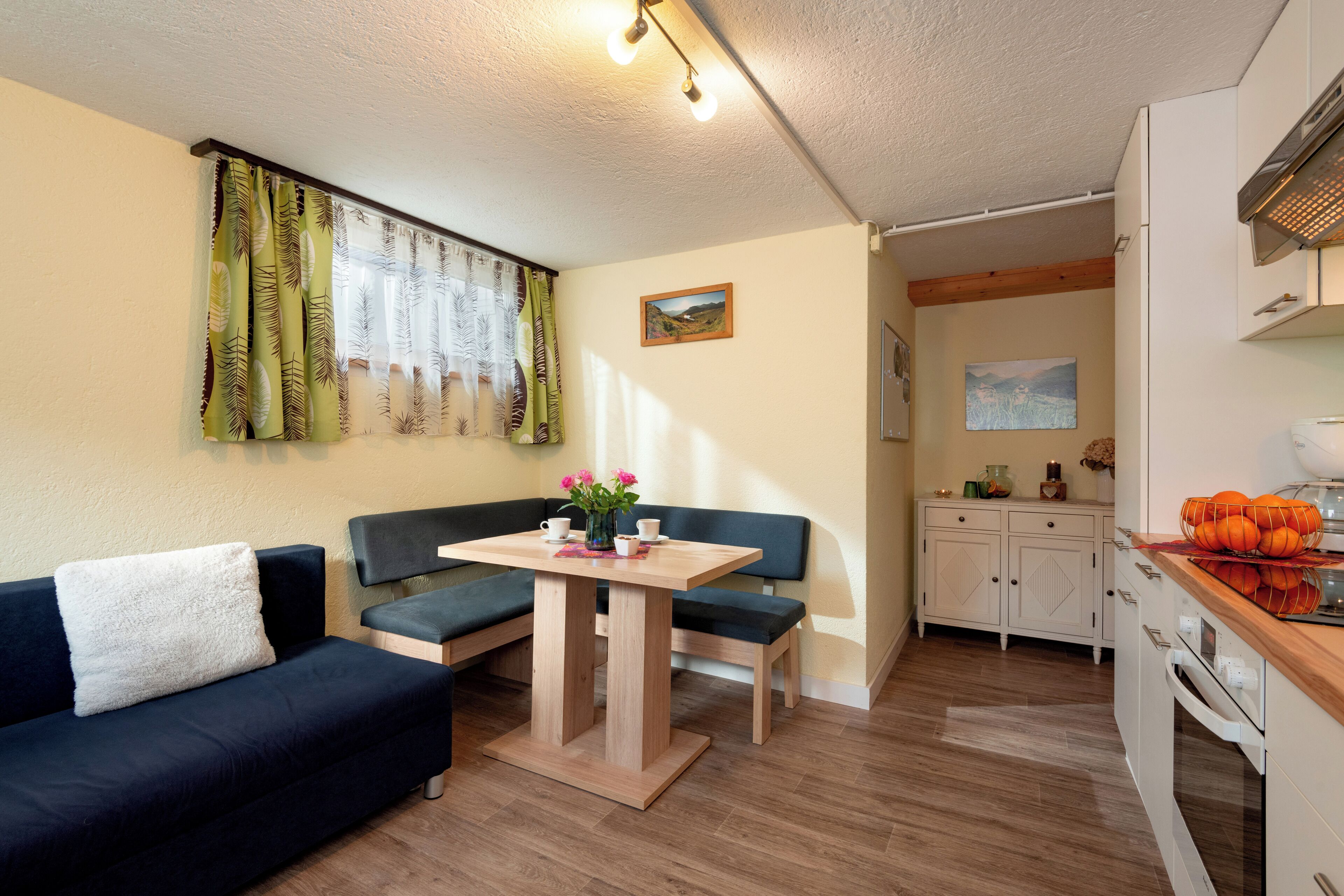 Un salon chaleureux avec un canapé bleu foncé, une table en bois avec deux chaises, et un coin cuisine doté de meubles blancs et d’un plan de travail en bois.