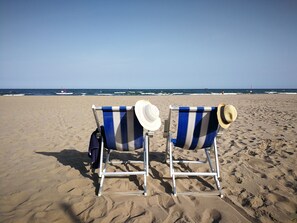 Deux chaises de plage aux coussins rayés bleu et blanc, ainsi que des chapeaux de paille, sont placés sur une plage de sable, d’où l’on peut admirer l’océan et apercevoir une bouée au loin.
