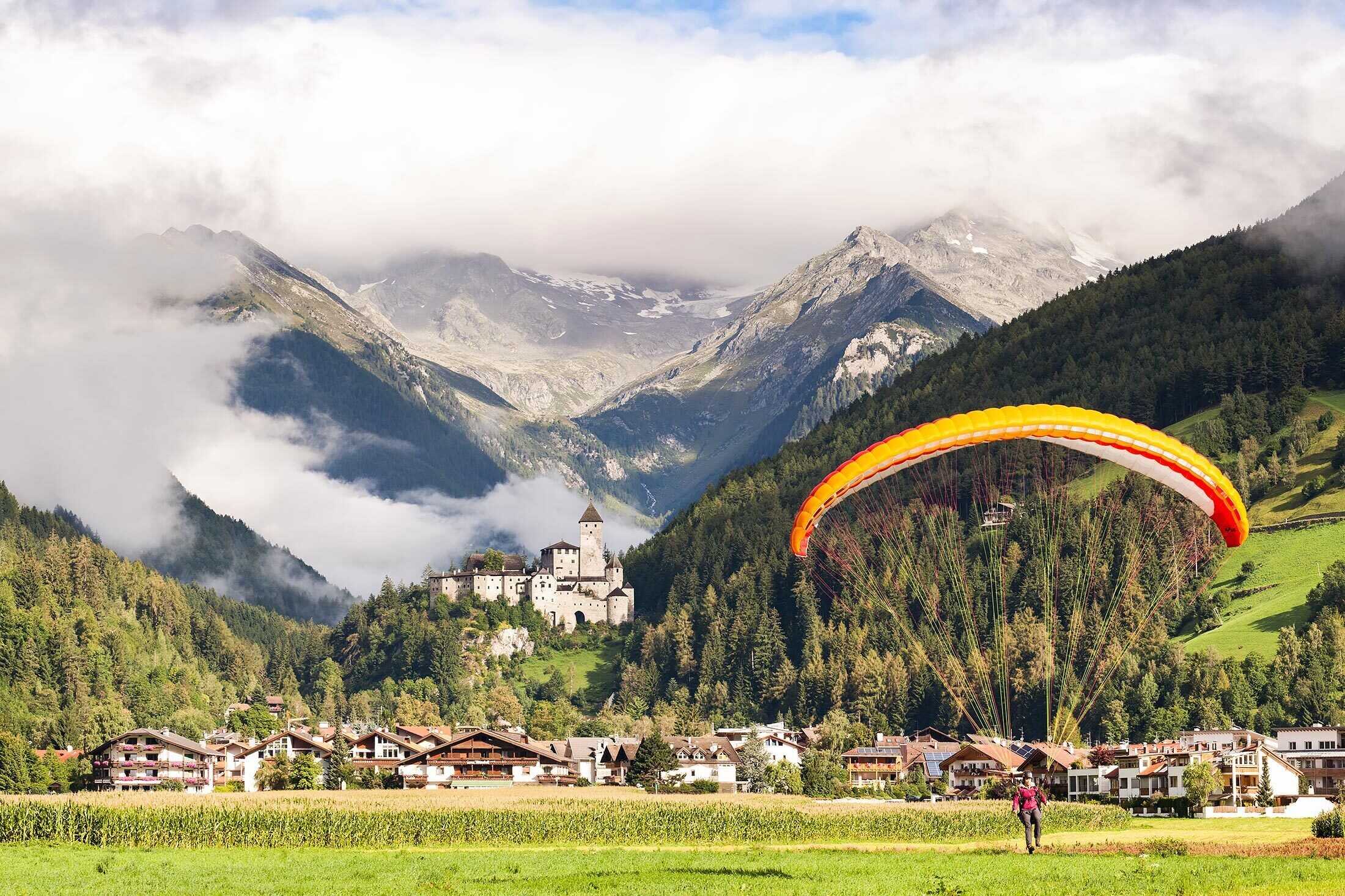 Un paisaje montañoso con una edificación que recuerda a un castillo, rodeado de abundante vegetación, y una persona practicando parapente.