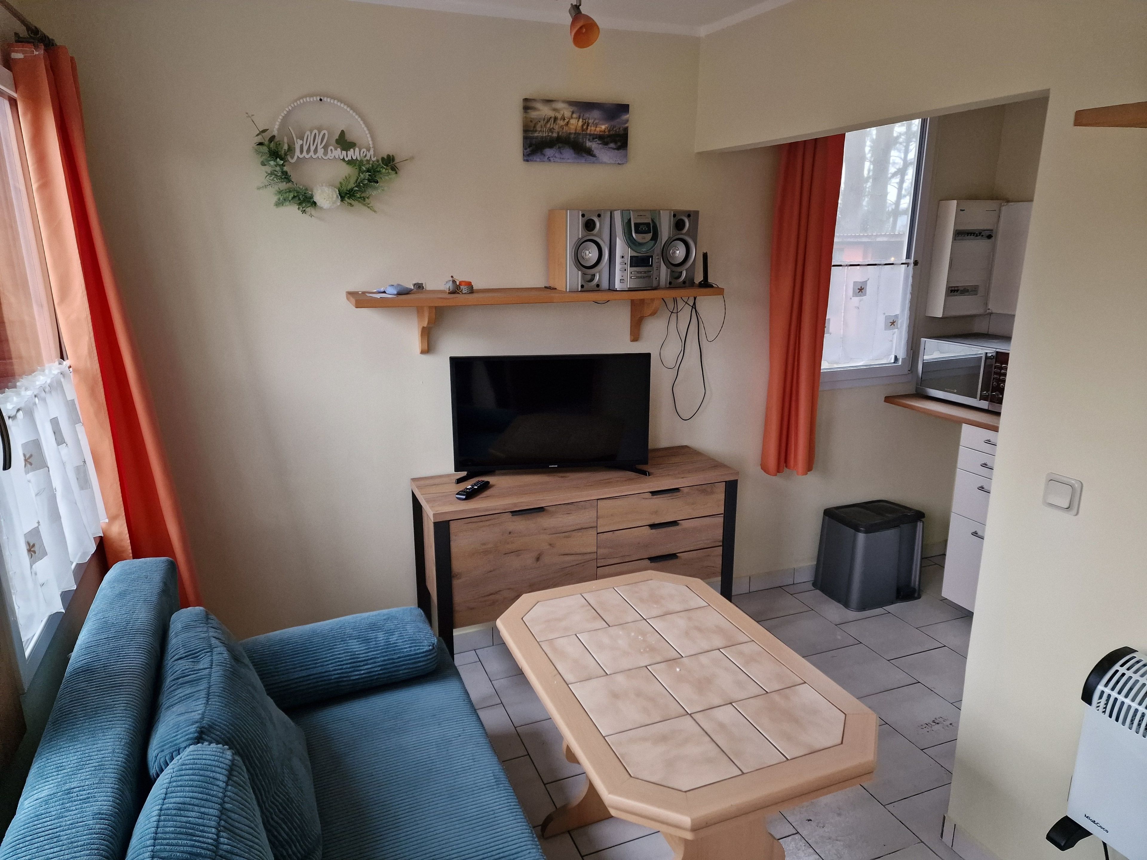 En kompakt opholdsstue med sofa, fjernsyn, lille bord og et tekøkken med mikrobølgeovn samt en skraldespand.