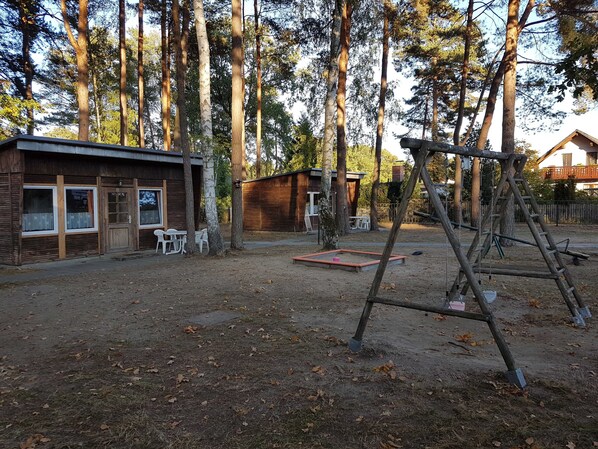 Un chalet rustique avec une façade en bois, un petit patio doté de meubles d’extérieur et une balançoire installée dans un espace boisé.