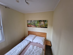 Et soveværelse med en træseng, hvide sengetæpper, et natbord, et billede af en skov og et vindue med dør.