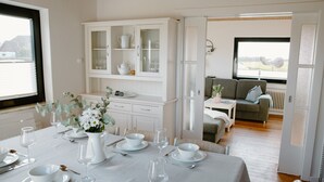 A dining area with a table set for four, a white cabinet with glass doors, and a view of the outdoors through a door. - Gemütl cottage + wifi, 1.5 km to the beach, fenced playground / lawn (Pommerby)