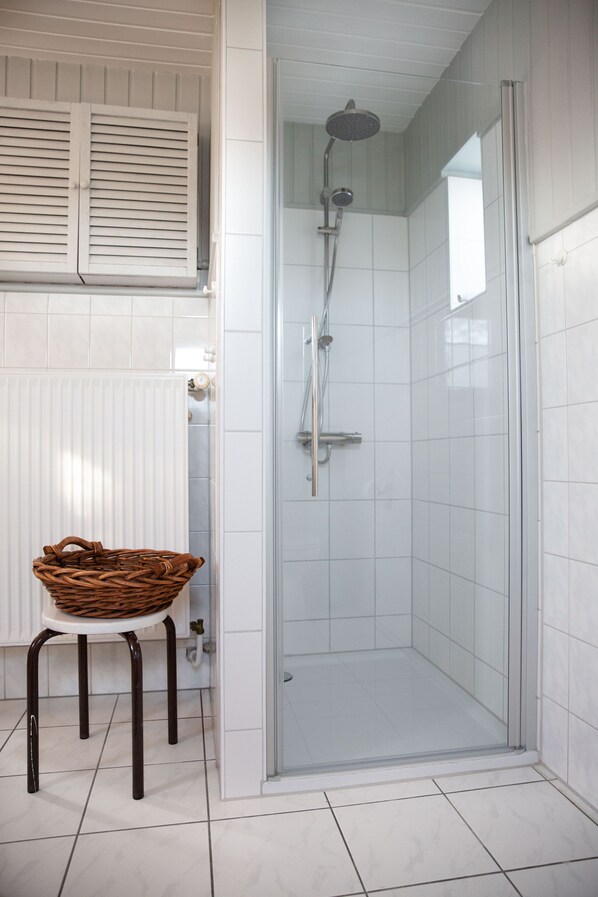 A glass-enclosed shower with a handheld showerhead, a white radiator, and a wicker basket on a stool.