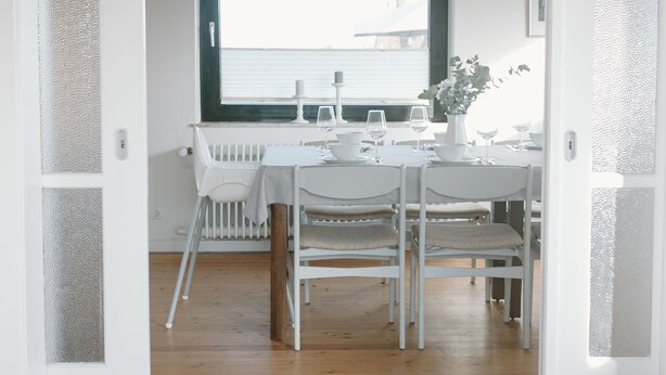 A dining area with a white table set for four, a window with a view, and a wooden ceiling. - Gemütl cottage + wifi, 1.5 km to the beach, fenced playground / lawn (Pommerby)
