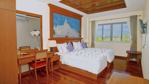 Une chambre d’hôtel avec un lit en bois, un bureau avec une chaise, un tableau représentant un temple, une fenêtre donnant sur un paysage verdoyant et un miroir.