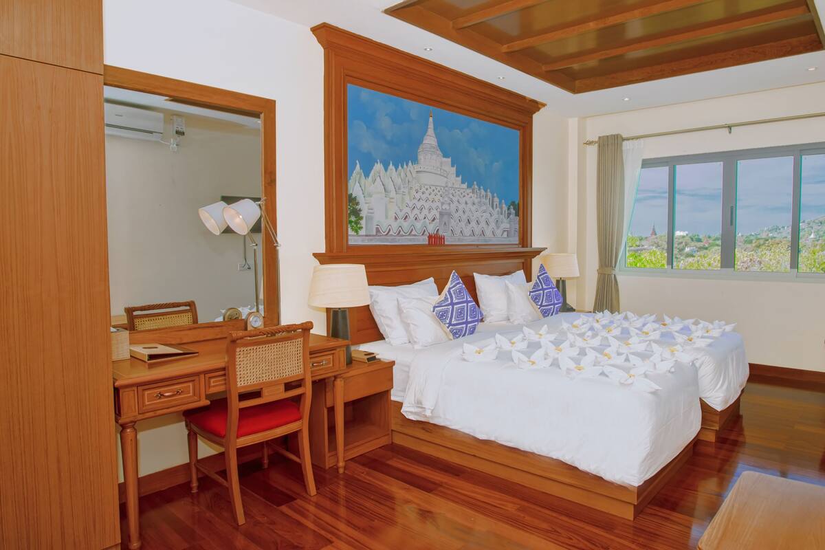 A hotel room with a wooden bed, a desk with a chair, a painting of a temple, a window with a view of greenery, and a mirror.
