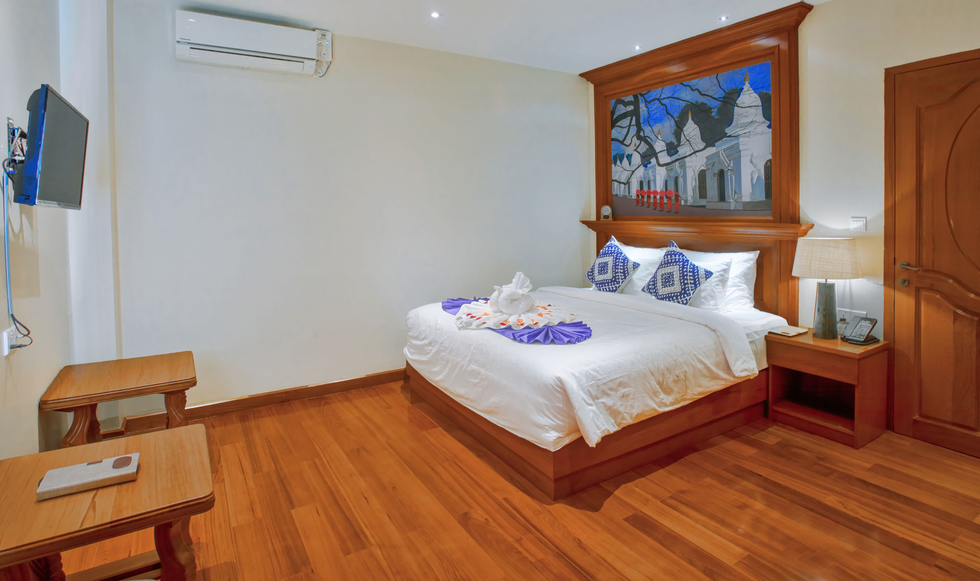 A hotel room with a wooden bed, a painting above the bed, a bedside table with a lamp, and a wooden nightstand.