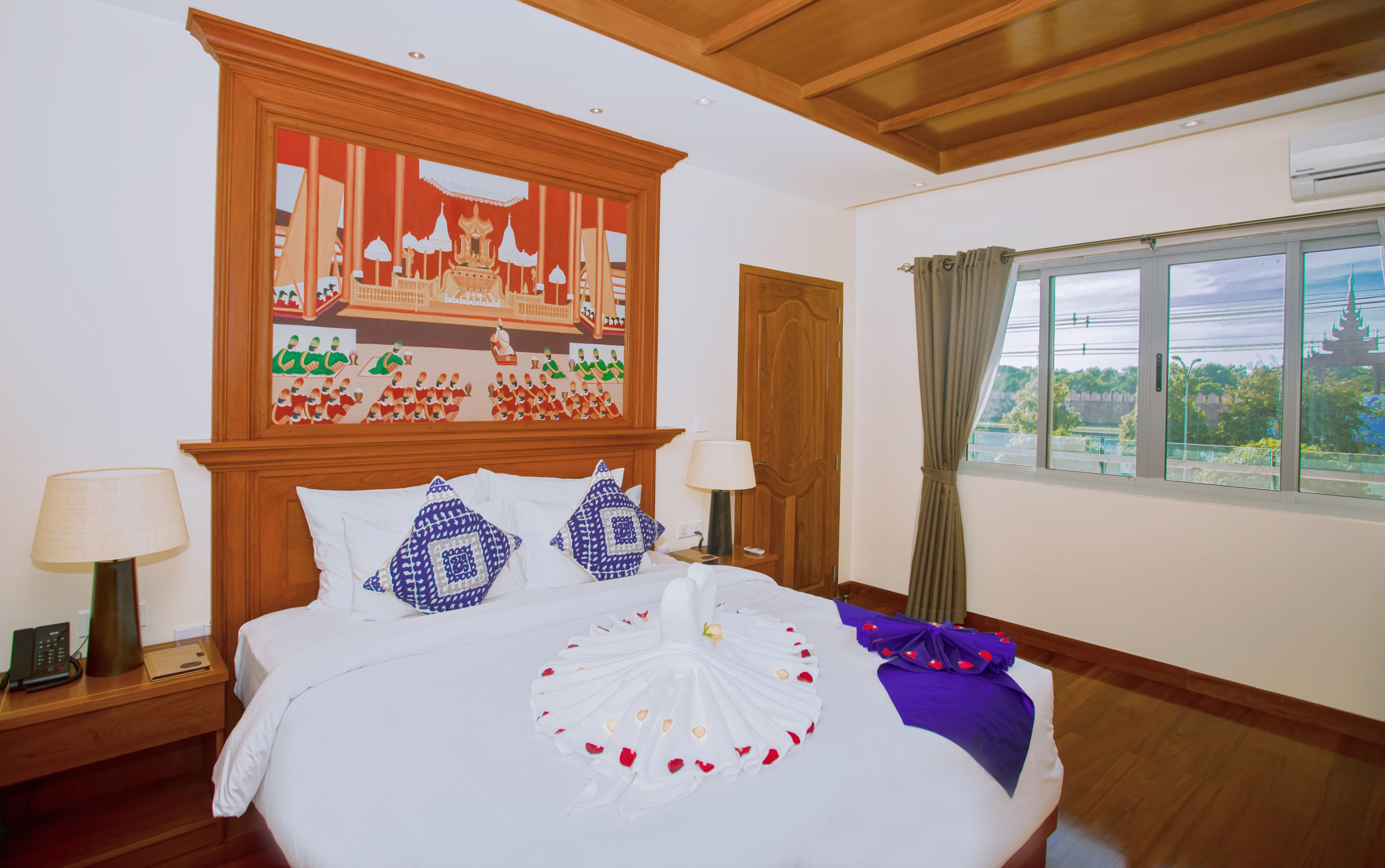 A hotel room with a large bed, wooden headboard, and a painting above it. There is a window with curtains and a view of greenery outside.
