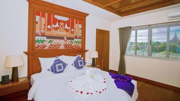 A hotel room with a large bed, wooden headboard, and a painting above it. There is a window with curtains and a view of greenery outside.