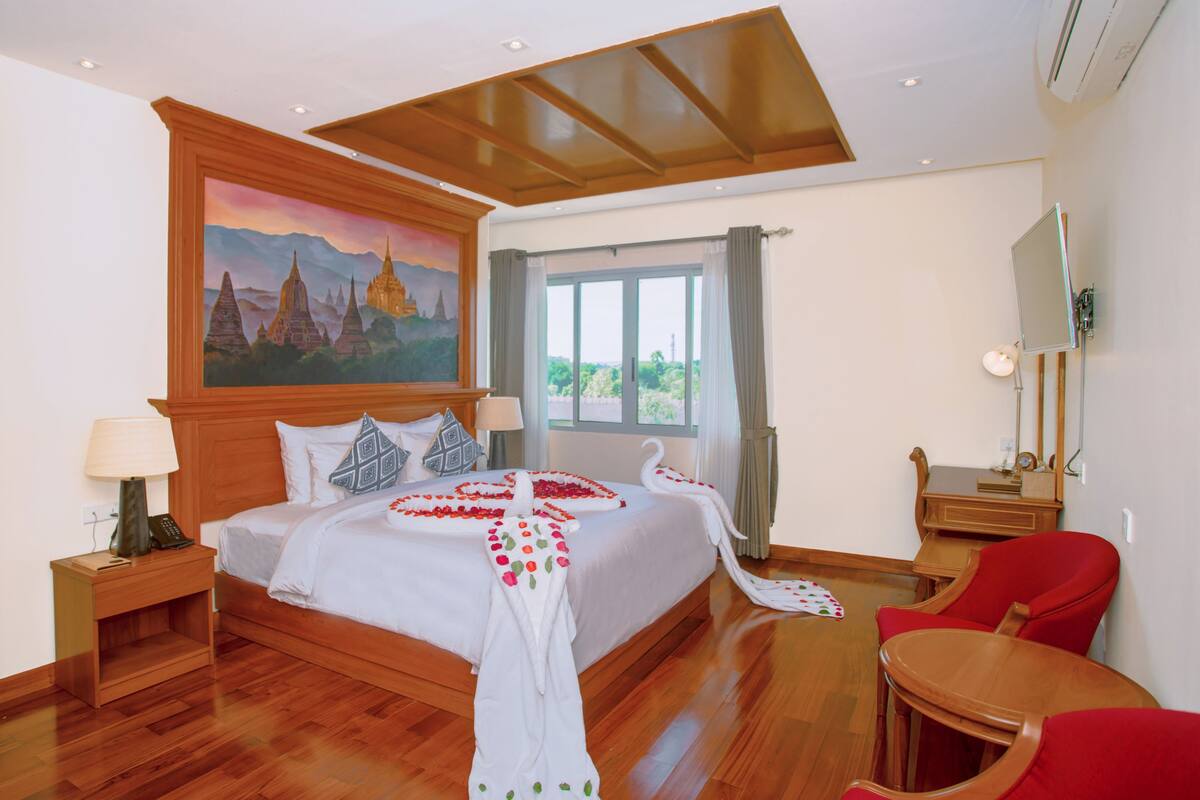 A hotel room with a large bed, a painting of a temple, a desk with a lamp, and a red chair.