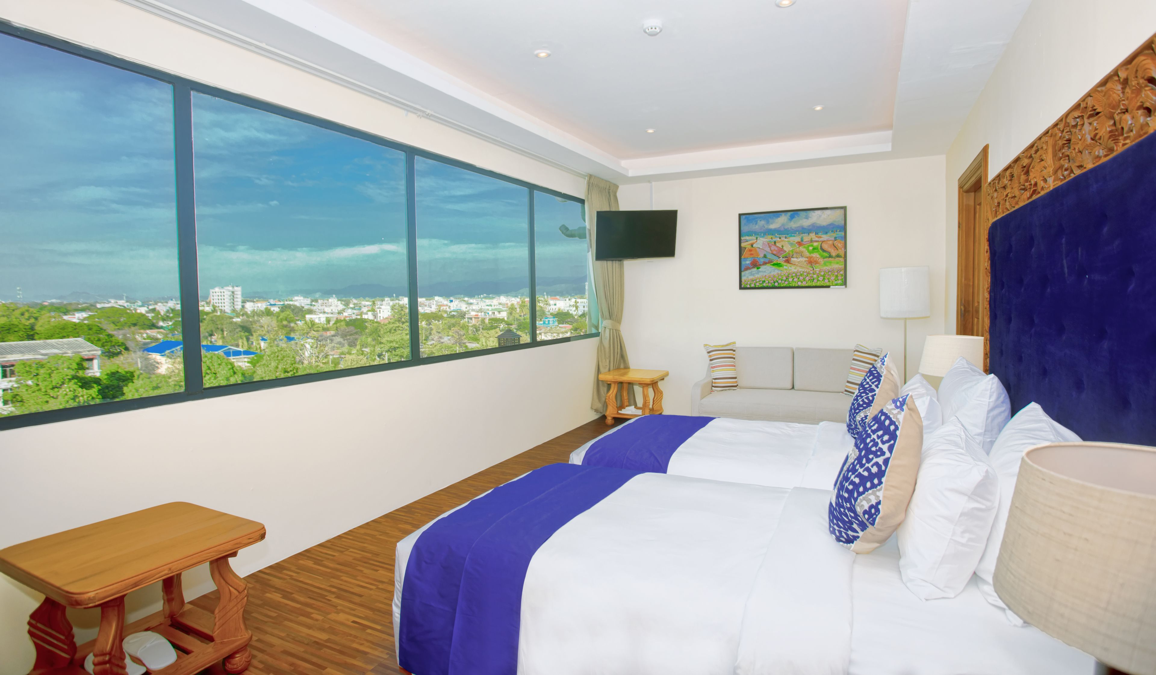 A hotel room with a large window, a bed with white and blue bedding, a wooden nightstand, a small table, a TV, and a painting on the wall.
