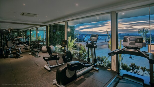 Une salle de sport équipée de divers appareils d’exercice, dotée de grandes fenêtres et offrant une vue sur le paysage urbain.