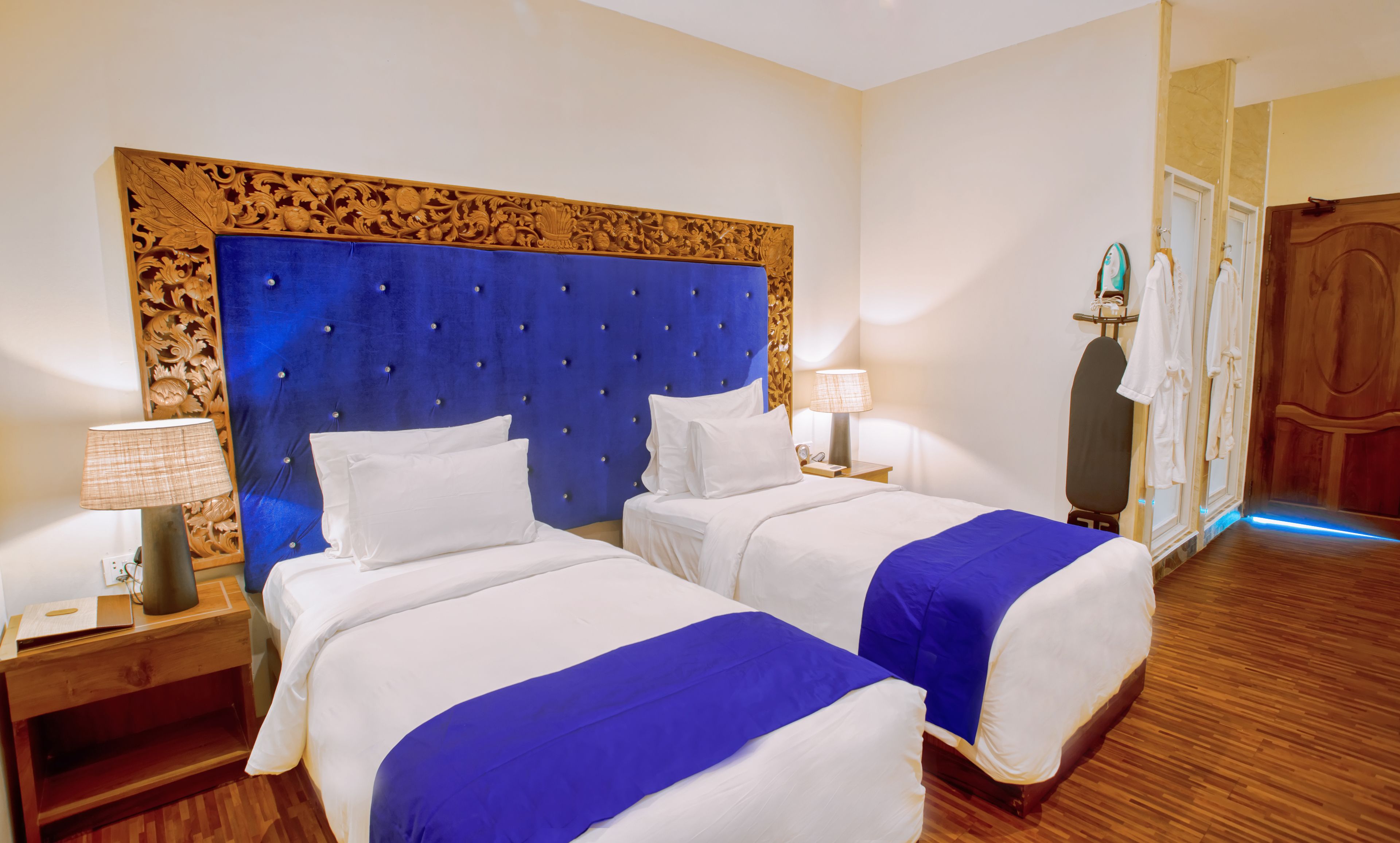 A hotel room with two beds, a wooden headboard, a nightstand with a lamp, and a wooden floor.