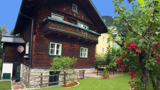 Una casa de madera tradicional con base de piedra, un balcón con detalles verdes y un jardín bien cuidado con plantas en flor.